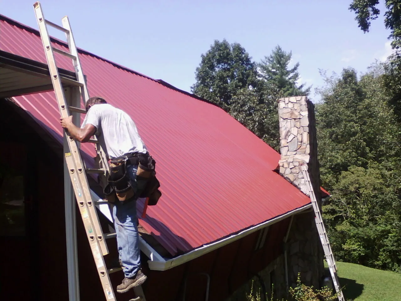 Roofer on a ladder beside a red metal roof and stone chimney — Soffit & Fascia Repair by Roofing Brothers in Spiceland